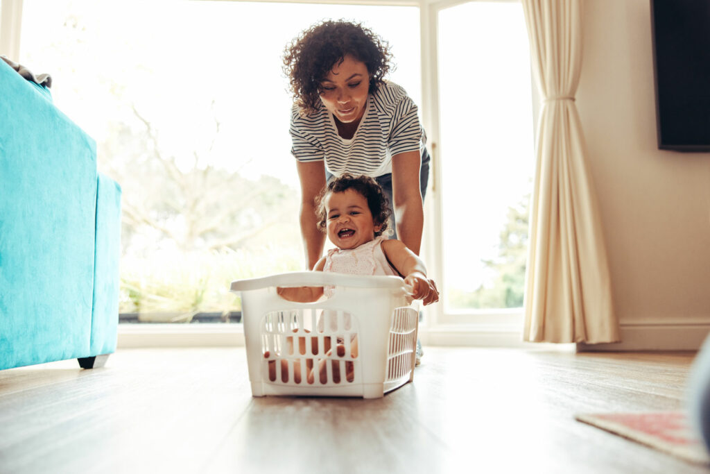 Woman pushing smiling baby sitting in washing basket. Baby enjoying laundry basket ride at home.